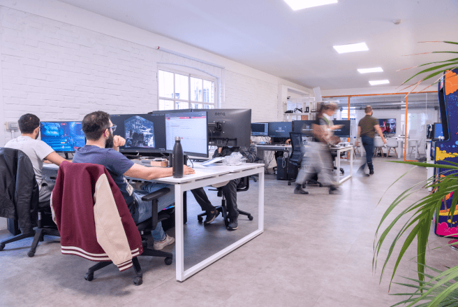 An open office space with team members working at desks with multiple monitors, and others walking in the background. An open office space with team members working at desks with multiple monitors, and others walking in the background.