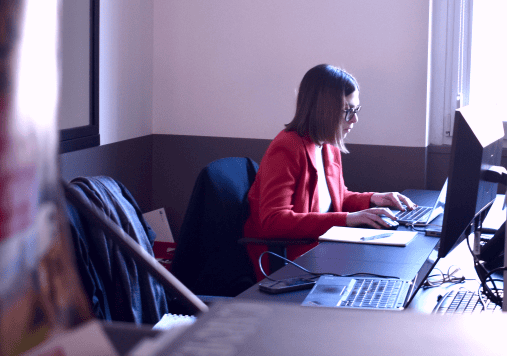A team member working at a desk in an office environment, focused on a computer screen, wearing a red blazer. A team member working at a desk in an office environment, focused on a computer screen, wearing a red blazer.