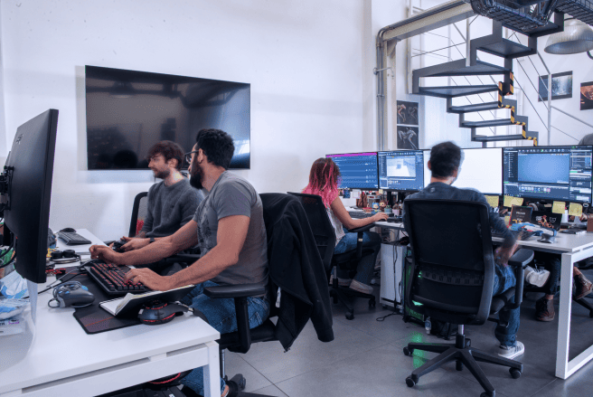 A group of team members working at desks in a modern office, with a staircase and a large screen in the background. A group of team members working at desks in a modern office, with a staircase and a large screen in the background.