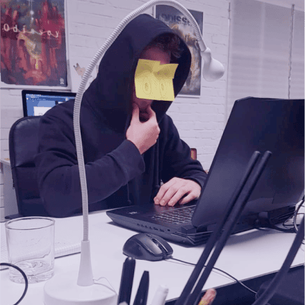 A person at a desk with a laptop, wearing a hoodie and holding a Post-it note with a drawing over their face. A person at a desk with a laptop, wearing a hoodie and holding a Post-it note with a drawing over their face.